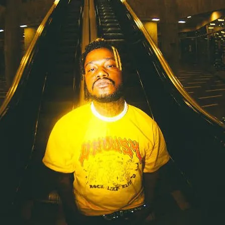 O rapper Bruno Sales, de camiseta amarela, em uma escada rolante iluminada